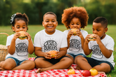 Four children sitting on a blanket outdoors, eating burgers with 'Jesus Burgers' shirts.